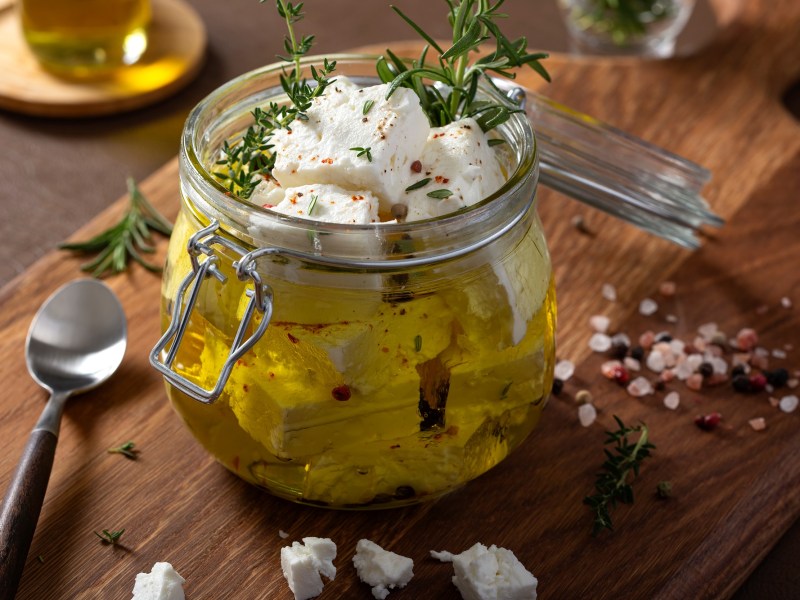 Glas mit mariniertem Feta auf einem Holzbrett