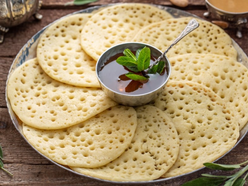 Einige Baghrir – marokkanische Pfannkuchen –, die auf im Kreis auf einem Teller angeordnet sind. In der Mitte steht eine Schale mit Honig-Butter-Soße.