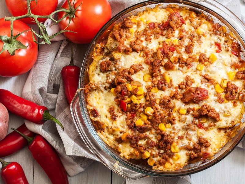 Mexikanischer Kartoffel-Hack-Auflauf in einer Auflaufform, daneben Tomaten, Chilis und Kartoffeln.