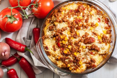 Kartoffel-Hack-Auflauf nach mexikanischer Art in einer Auflaufform, daneben Tomaten, Chilis und Kartoffeln.