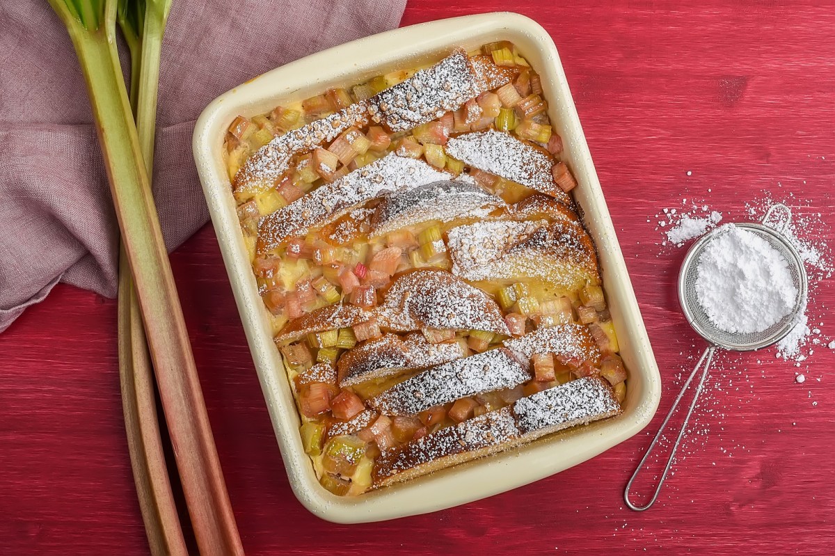 Auflaufform mit Brioche-Auflauf mit Rhabarber auf rotem Untergrund. Puderzucker und Rhabarber daneben.