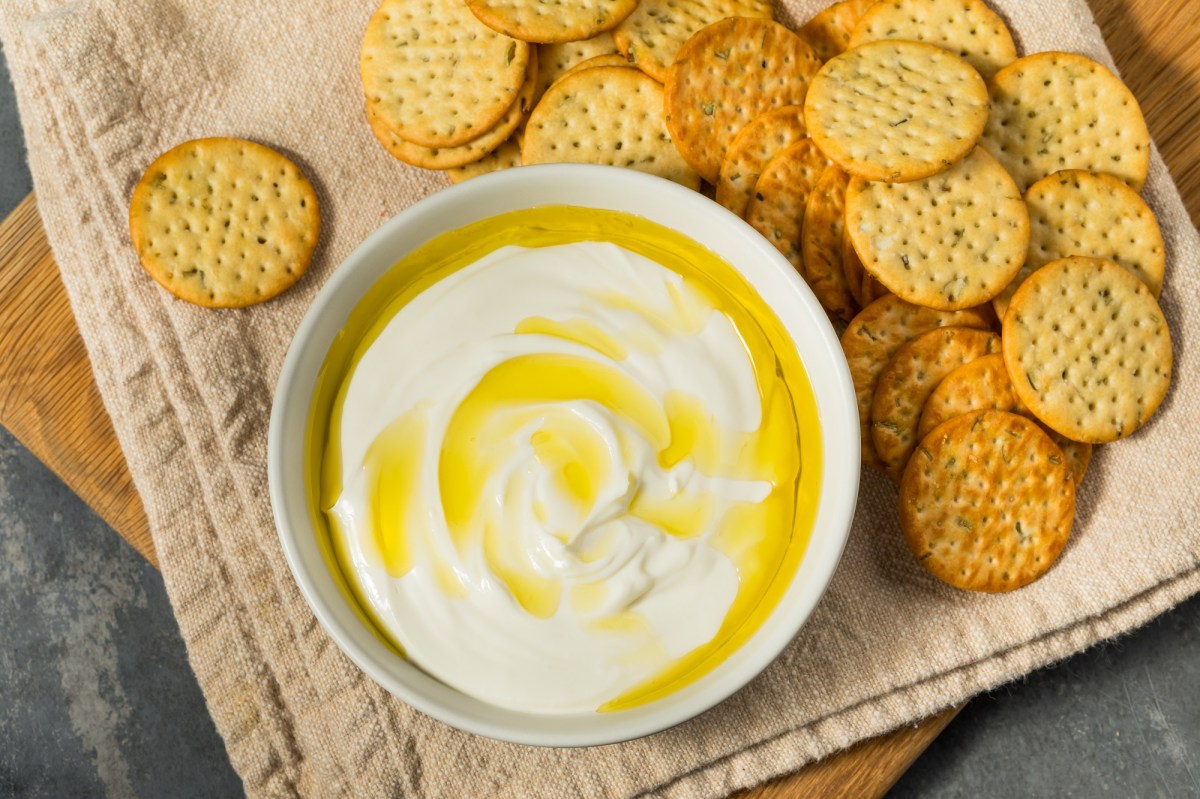 Ricotta-Dip mit Olivenöl neben einigen Crackern auf einem Holzbrett.