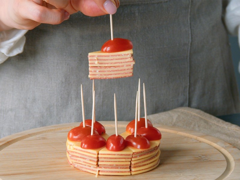 Salami-Käse-Snack mit Tomate auf einem Holzbrett. Die Hand einer Köchin hält ein Stück in die Kamera