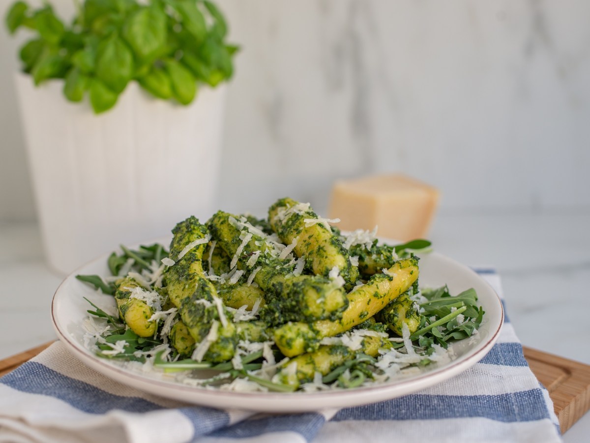 Ein Teller Schupfnudelpfanne mit Spargel und Rucola-Pesto mit Parmesan bestreut steht auf einem blau-weiß-gestreiften Handtuch.