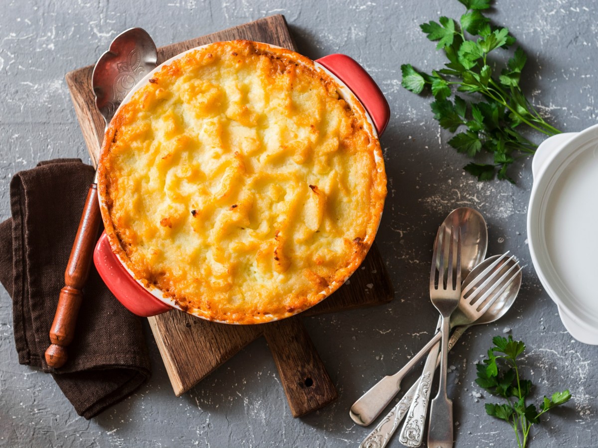 Shepherd's Pie mit Linsen und Kartoffelpüree in runder Auflaufform in der Draufsicht, daneben Besteck und Kräuter.