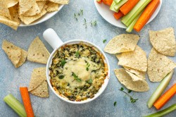 Spinat-Artischocken-Dip in einer weißen Schale. Daneben Tortillachips und Gemüsesticks