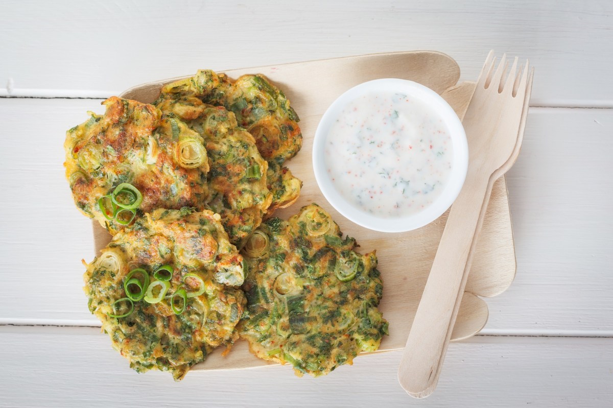 Zucchinipuffer auf einer Pappunterlage. Daneben ein Dip und eine Holzgabel.