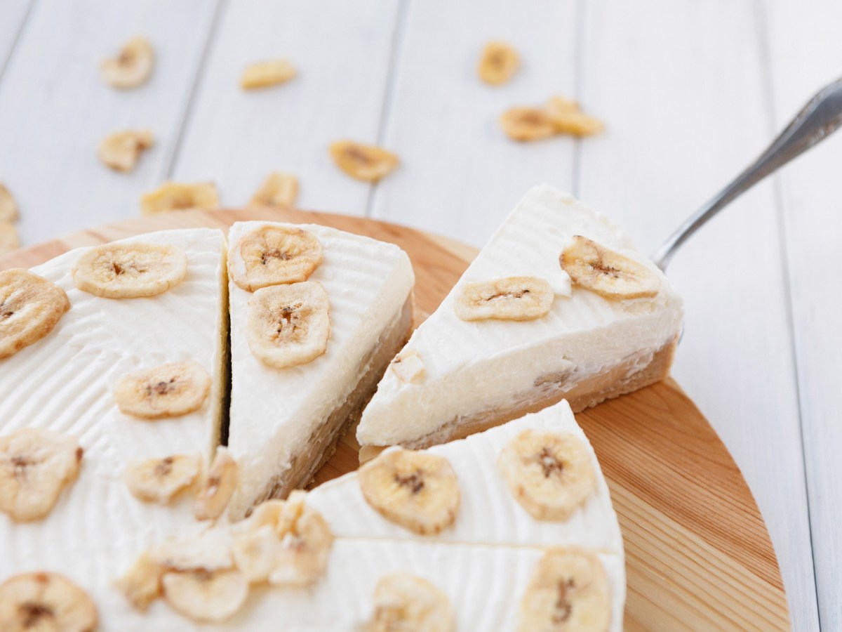 Bananen-Mascarpone-Torte auf einem Holzbrett. Mit einem Kuchenheber wird ein Stück entnommen.