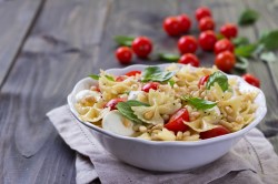 Eine weiße Schüssel Caprese-Nudelsalat mit Tomaten, Mozzarella, Basilikum und Pinienkernen. Im Hintergrund ganze Tomaten am Strauch.