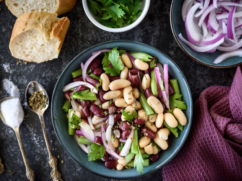 Schüssel mit Drei-Bohnen-Salat auf dunklem Tisch, Besteck und Baguette daneben.