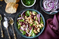 Schüssel mit Drei-Bohnen-Salat auf dunklem Tisch, Besteck und Baguette daneben.