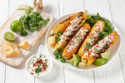 Elote - mexikanischer Streetfood-Mais auf einem weißen Teller. Daneben Limetten, Koriander und Käse.