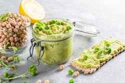 Erbsen-Hummus im Glas garniert mit Kresse, daneben Kichererbsen im Glas, eine halbe Zitrone und ein Knäckebrot mit Hummus bestrichen.