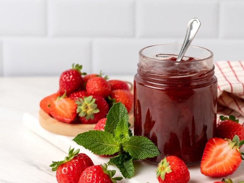 Ein Glas Erdbeer-Kokos-Marmelade mit einem Löffel darin, drumherum frische Erdbeeren.