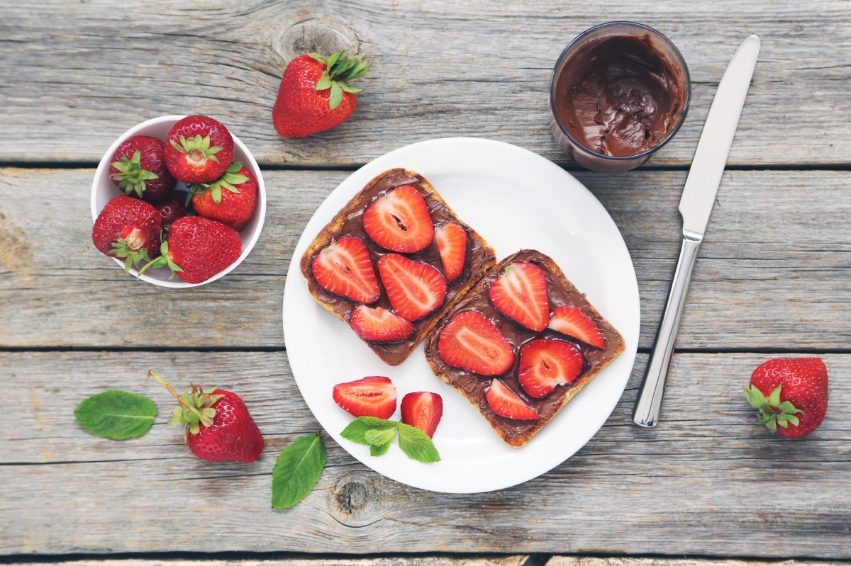 Erdbeer-Nutella-Toasts auf einem weoßen Teller. Daneben stehen ein Glas Nutella und es liegen einige Erdbeeren bereit.