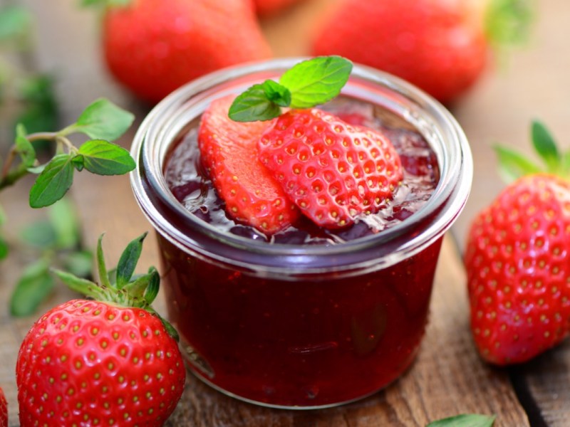 Ein Glas Erdbeermarmelade mit Minze und frischen Erdbeeren on top. Drumherum liegen Erdbeeren.