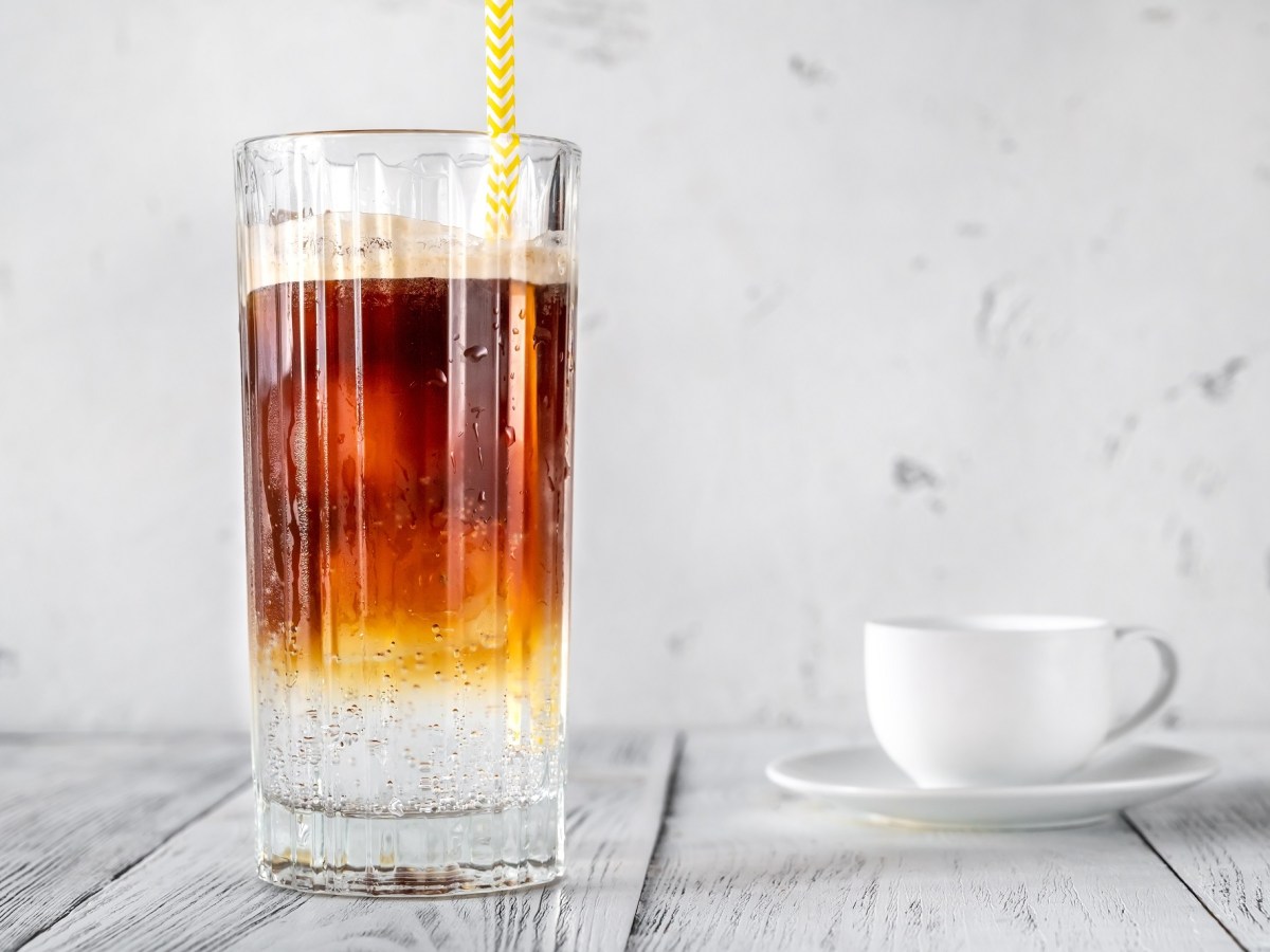 Ein Glas Espresso Tonic mit Eis und Strohhalm, daneben eine weiße Tasse mit Untertasse.