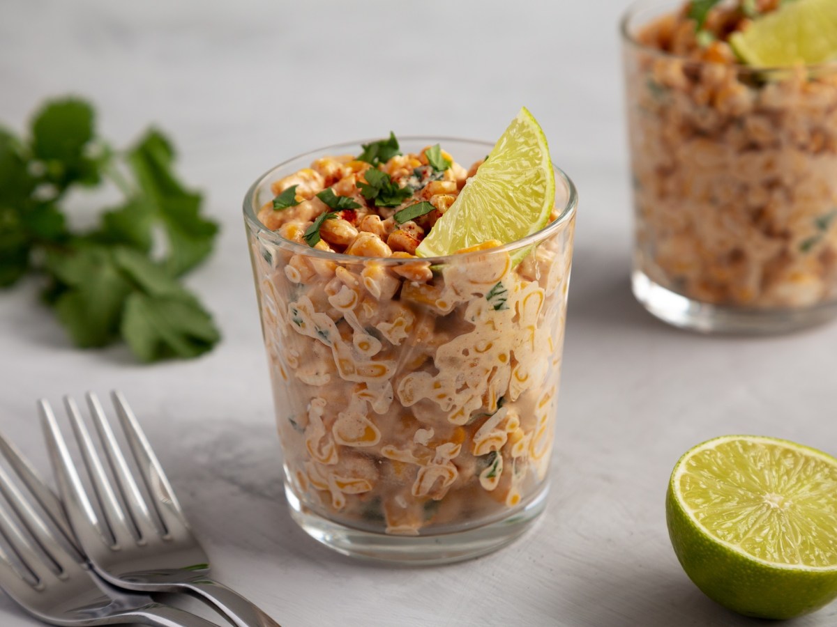 2 Gläser Esquites, mexikanischer Maissalat, garniert mit Limettenscheiben und Koriander, daneben zwei Gabeln, frischer Koriander und eine halbierte Limette.