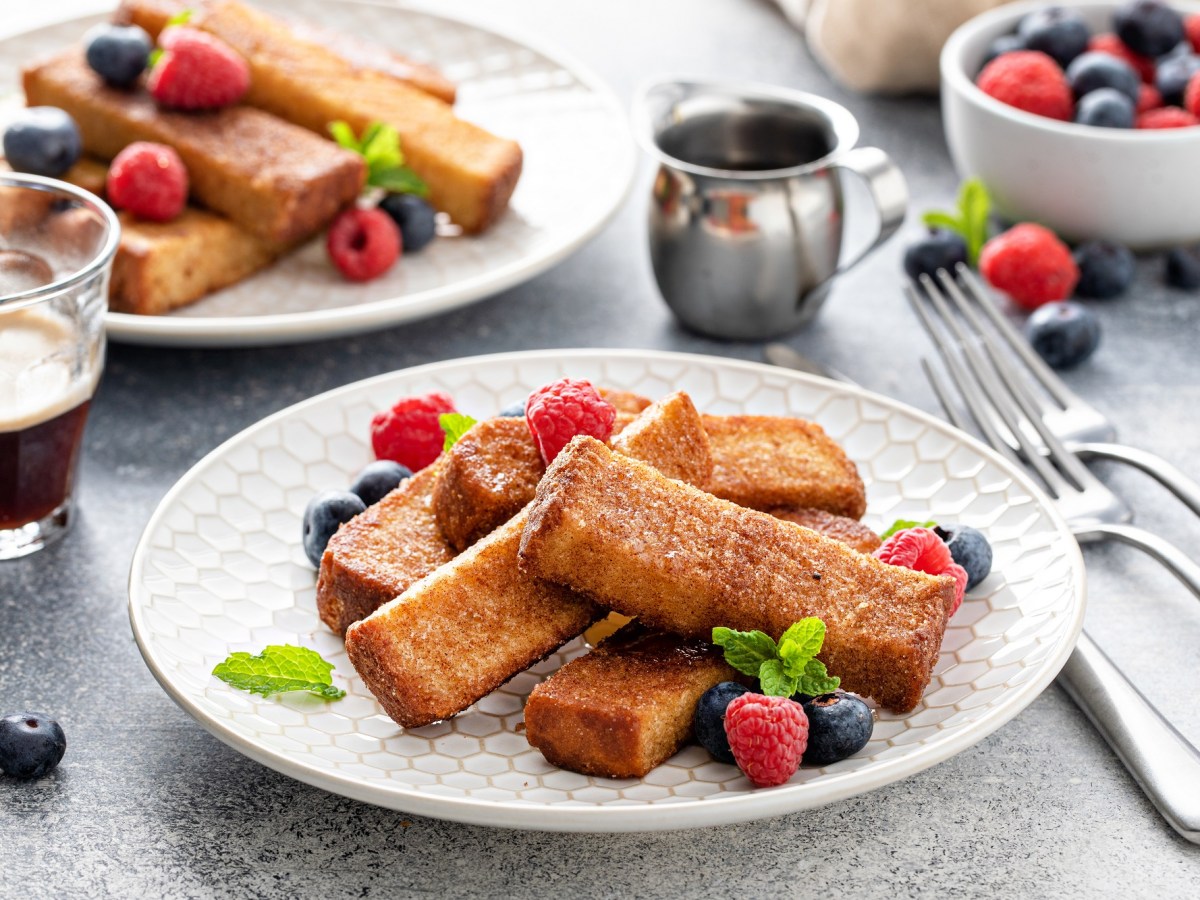 French-Toast-Stangen auf zwei Tellern, garniert mit Beeren, daneben Besteck.