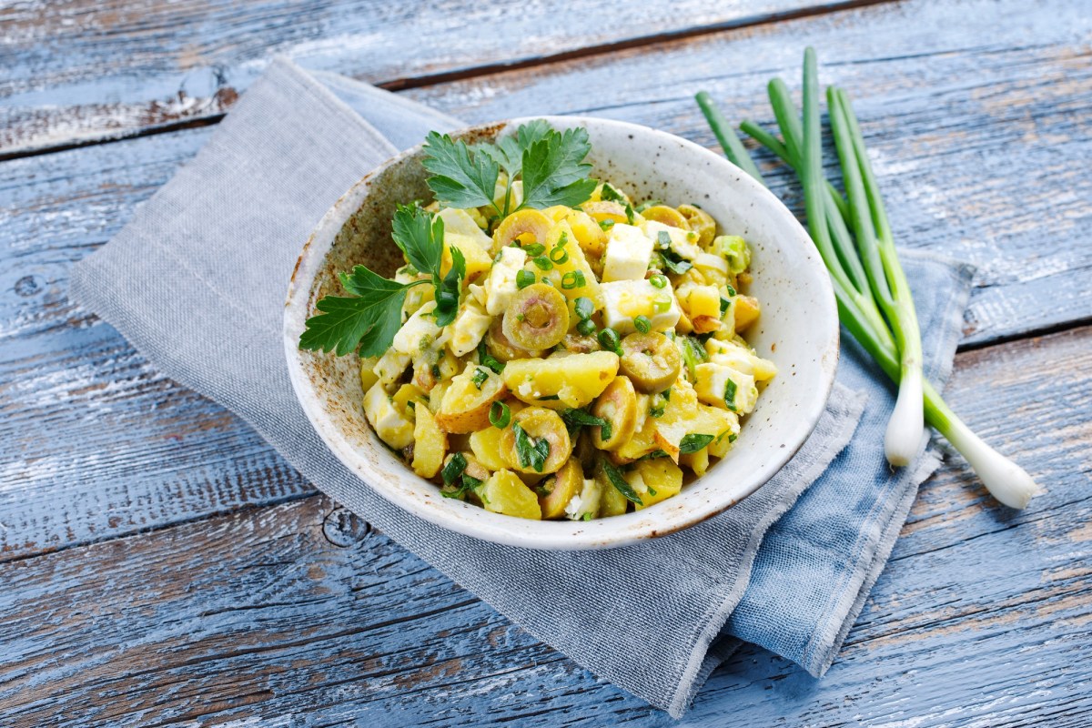 Griechischer Kartoffelsalat mit Oliven und Feta sowie Dill und Frühlingszwiebeln in einer Schüssel, die auf einer Stoffserviette liegt, daneben frische Frühlingszwiebeln.