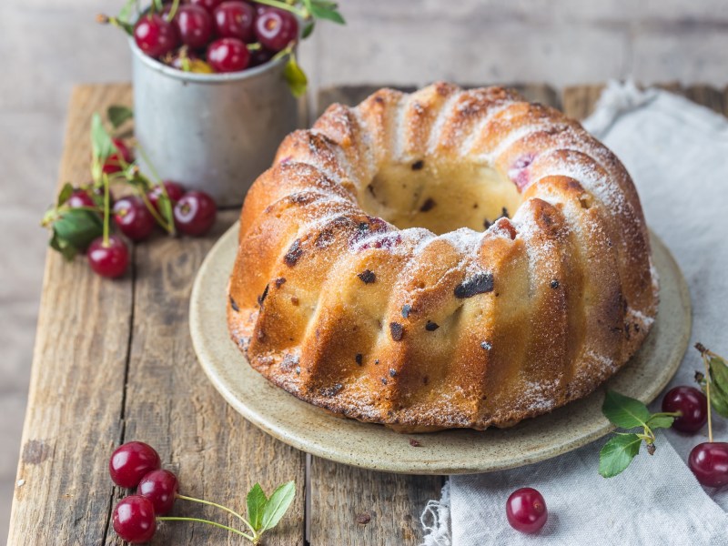 Ein Gugelhupf mit Kirschen auf einem Holztisch mit frischen Kirschen.