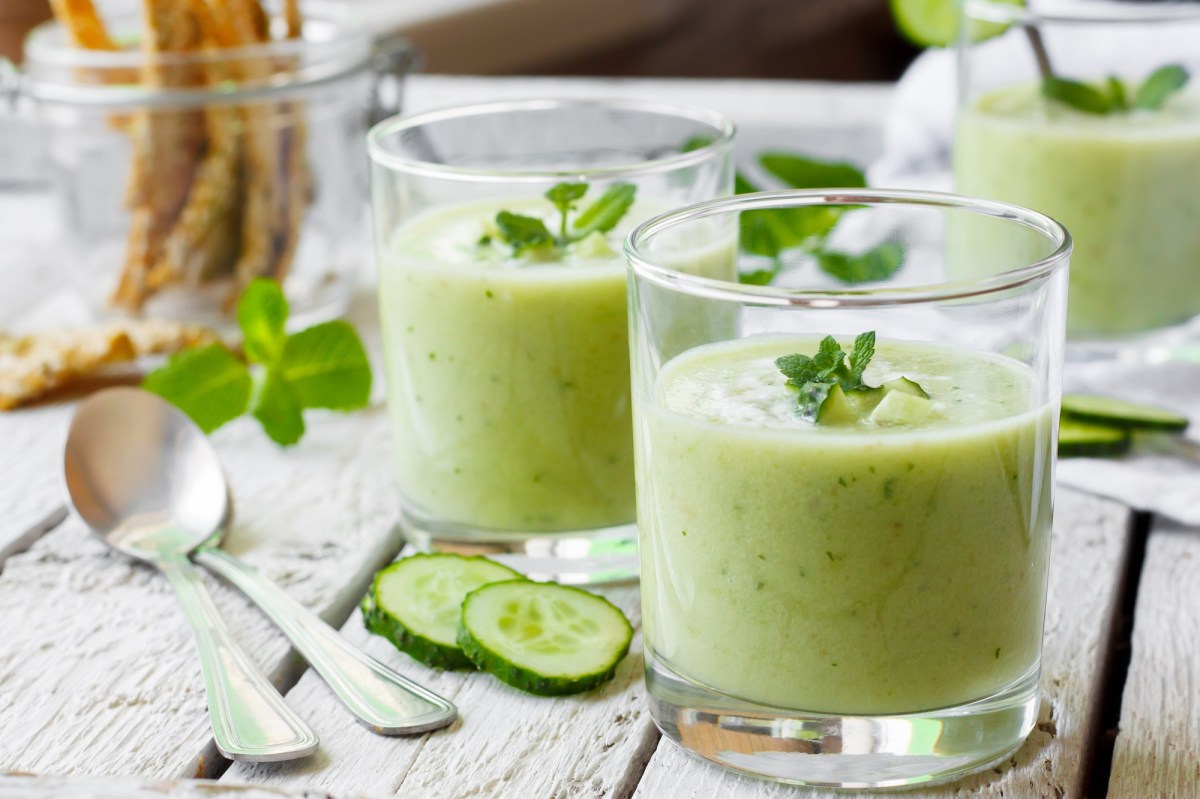 2 Gläser Gurken-Gazpacho garniert mit Minze, daneben zwei Löfel und Gurkenscheiben.