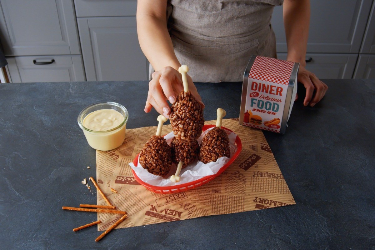 Eine Knusperkeule aus Reiscrispies wird von einer Hand gehalten. Weitere Keulen sind im Hintergrund zu sehen.