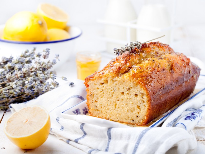 ein angeschnittener französischer Lavendelkuchen auf einem karierten Geschirrtuch, daneben Lavendel und Zitronen.