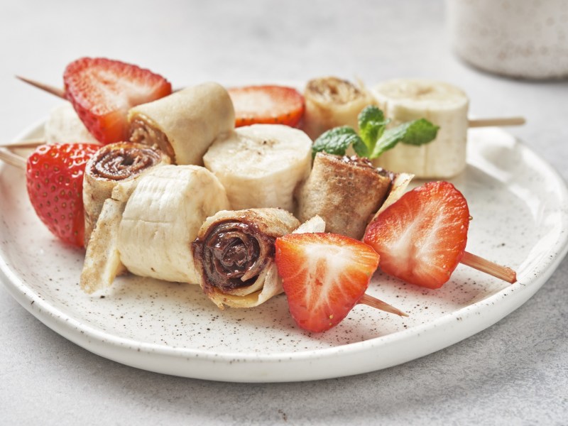 3 Pfannkuchen-Spieße mit Erdbeeren und Bananen auf einem Teller.