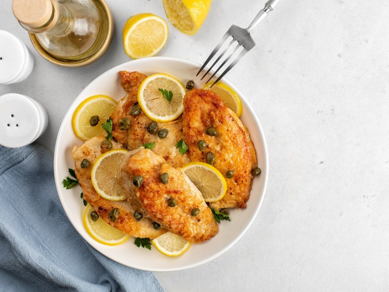 Piccata mit Hähnchen auf einem weißen Teler. Daneben liegt Zitrone und steht eine Flasche Olivenöl.