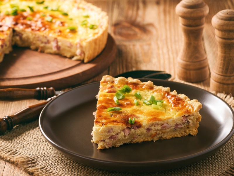 Ein Stücke Quiche Lorraine auf einem dunklen Teller, bestreut mir Frühlingszwiebeln. Im Hintergrund der Rest der Quiche