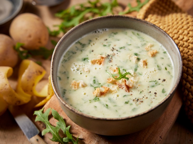 Eine Schüssel Rucola-Kartoffel-Suppe mit Croûtons, daneben Kartoffeln, Nahaufnahme.