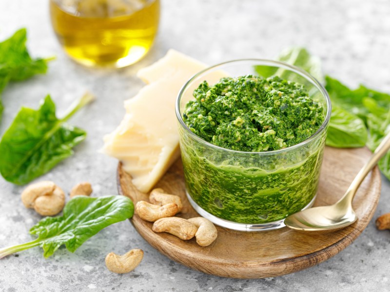 Ein Glas Spinatpesto auf einem runden Holzteller, drumherum frischer Spinat, Cashews und außerdem ein Glas Olivenöl.