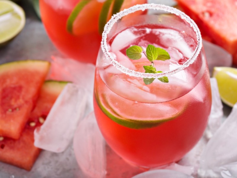 Ein Glas Watermelon Man. Drumherum liegen Limetten, Eiswürfel und frische Wassermelone.