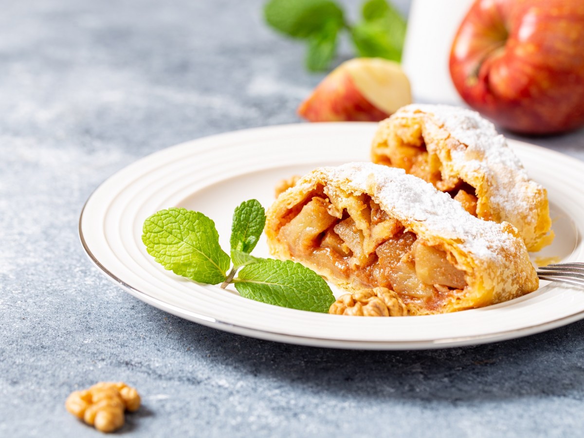 Zwei Stücke Apfelstrudel mit Blätterteig auf einem Teller, bestäubt mit Puderzucker, im Hintergrund Äpfel.