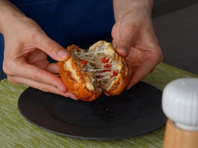 Eine Frau teilt ein gefülltes Hackfleisch-Käsebrötchen, man sieht nur ihre Hände, Nahaufnahme.