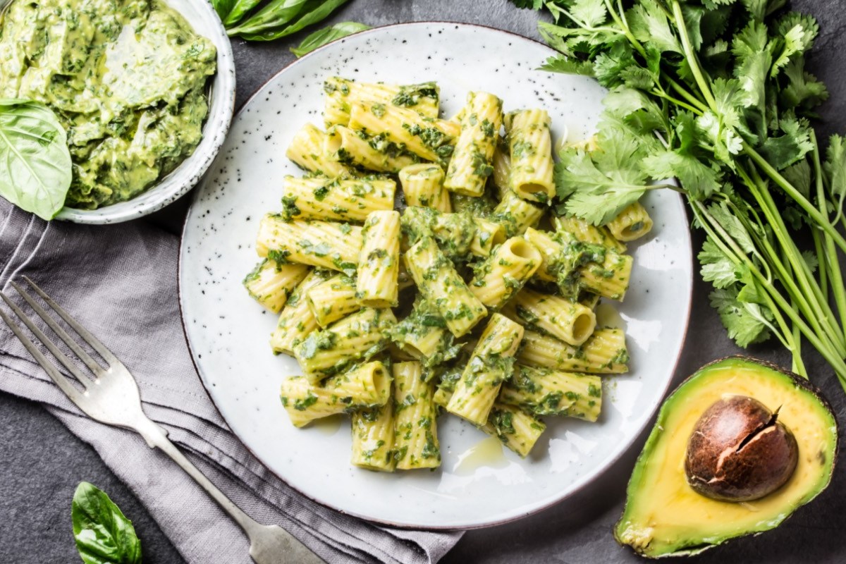 Ein Teller Nudeln mit Avocado-Pesto in der Draufsicht. Daneben eine Schüssel Pesto und frische Zutaten.