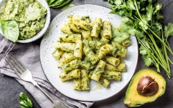 Ein Teller Nudeln mit Avocado-Pesto in der Draufsicht. Daneben eine Schüssel Pesto und frische Zutaten.