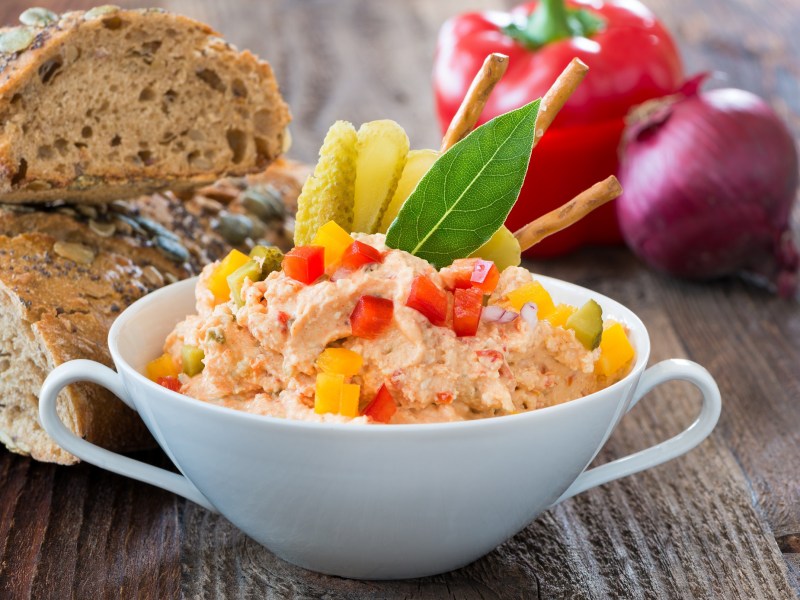 Paprika-Aufstrich mit Frischkäse in einer Tasse, mit Paprikastücken, im Hintergrund Brot und Paprika und eine Zwiebel.