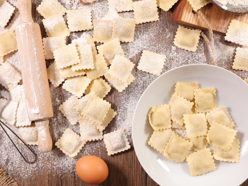 Ravioli selber machen: Selbstgemachte Ravioli auf einer Arbeitsfläche. Daneben liegen die Zutaten wie Ei und Mehl, sowie ein Nudelholz.