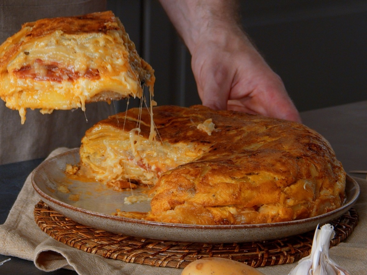 Spanische Tortilla auf einem braunen Teller. Eine männliche Hand hebt gerade ein Stück hoch, von dem sich Käsefäden zum Rest der Tortilla ziehen.