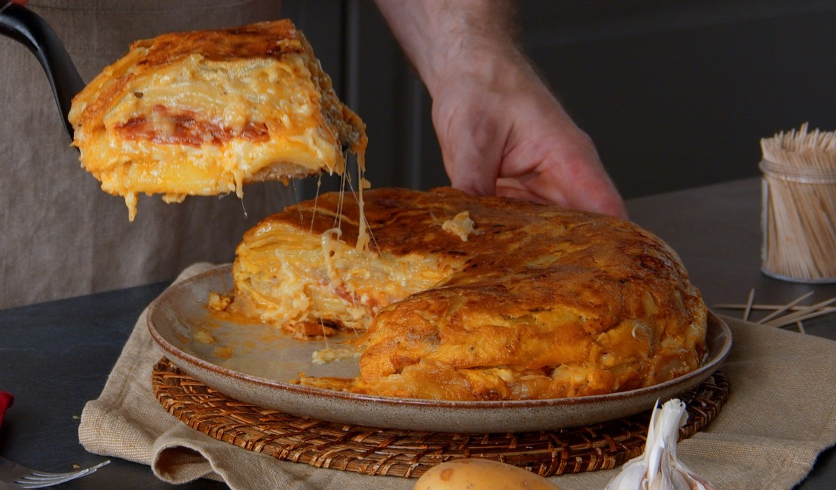 Spanische Tortilla auf einem braunen Teller. Eine männliche Hand hebt gerade ein Stück hoch, von dem sich Käsefäden zum Rest der Tortilla ziehen.