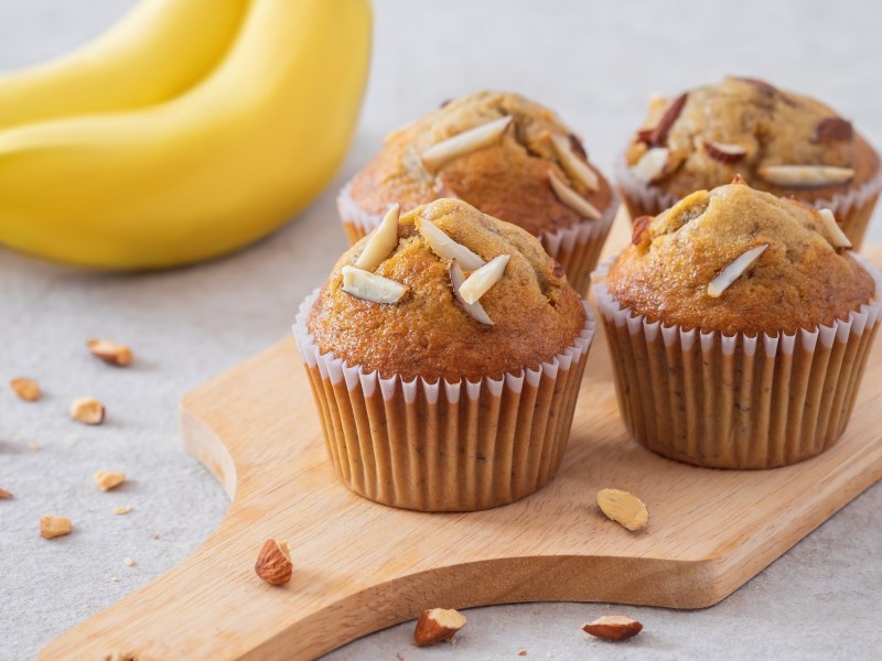 4 Bananenmuffins ohne Zucker auf einem Brett, garniert mit Mandelstiften, dahinter Bananen.