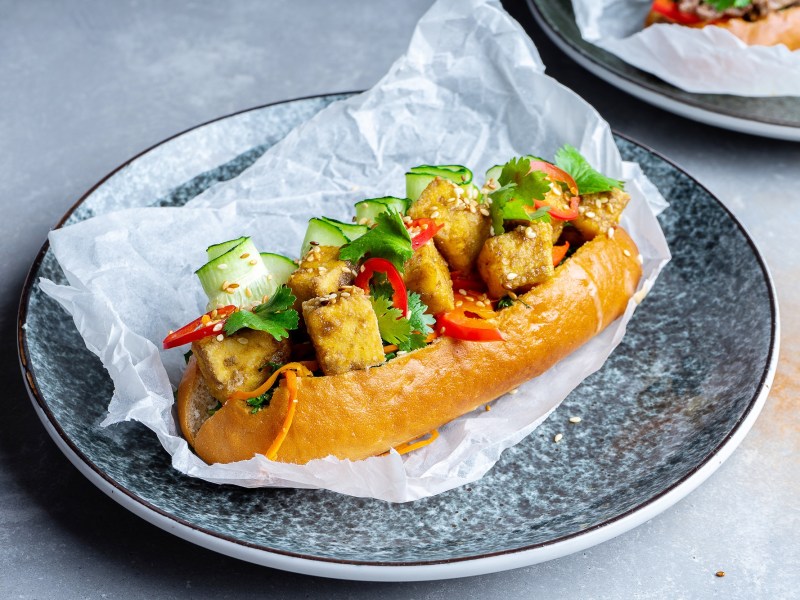 Ein Banh Mi Sandwich mit Tofu, Gurke, Paprika Koriander auf einem Teller.