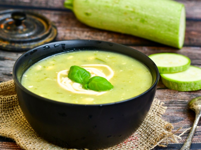 Eine Schüssel gegrillte Zucchinisuppe, garniert mit Basilikum, dahinter eine angeschnittene Zucchini.