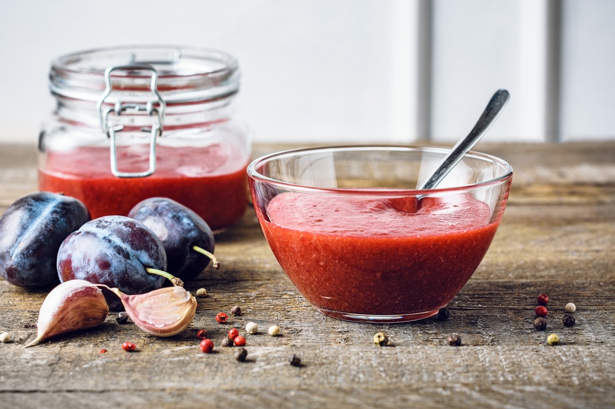 Pflaumenketchup in einer Schale. Daneben liegen Pflaumen und Knoblauchzehen. Im Hintergrund steht ein Glas mit mehr Ketchup.