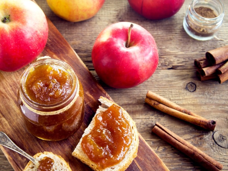 Ein Glas Bratapfelmarmelade. Daneben frische Äpfel und ein Brot mit Marmelade bestrichen.