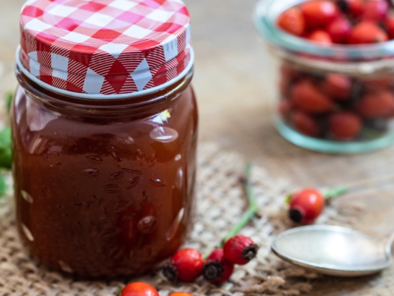 Ein Glas Hagebuttenmarmelade. Daneben frische Hagebutten in einer Glasschale.