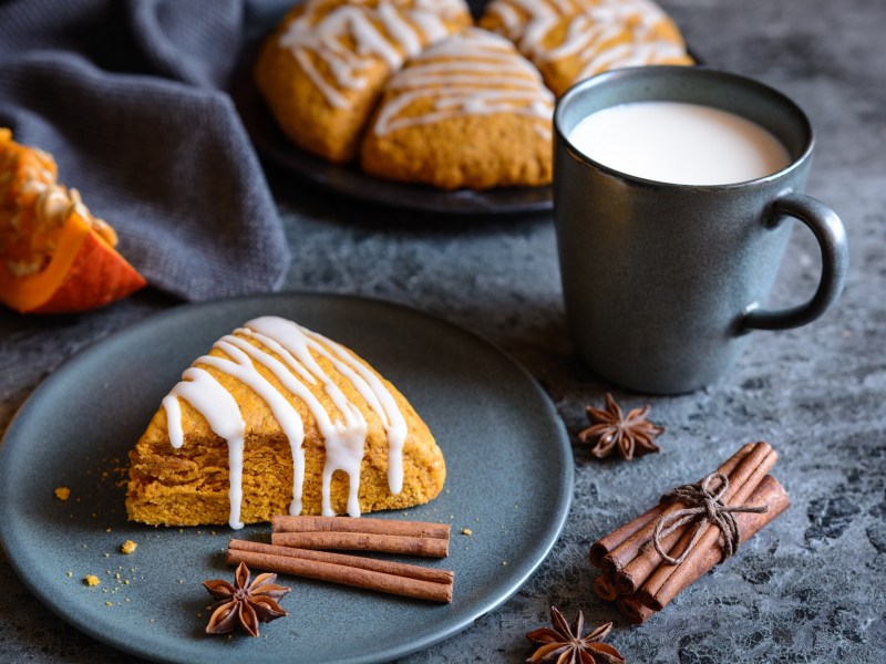 Ein Kürbis-Scones verziert mit Zuckerguss auf einem Teller, daneben Zimtstangen und eine Tasse Milch, im Hintergrund weitere Scones und Kürbisspalten.