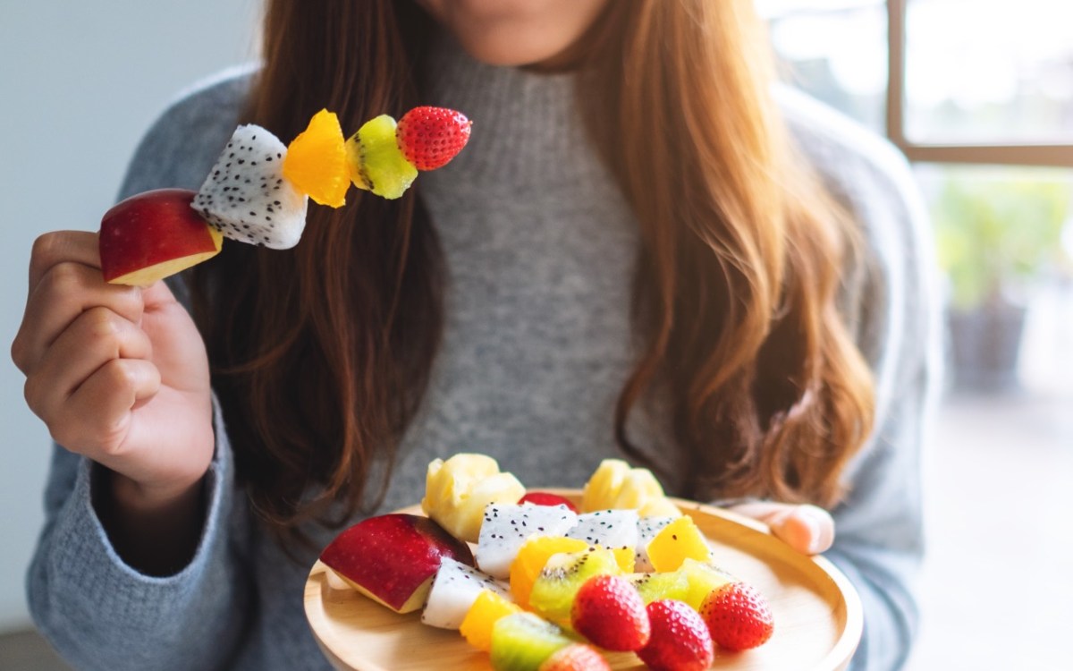 Obst am Abend essen: Eine Frau hält einen Obstspieß in der rechten und einen Teller mit Obstspießen in de rlinken Hand.